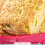 Close-up of golden brown Hatch Cheddar Biscuits with bits of green pepper, stacked in a pink basket. Text overlay reads "hatch cheddar biscuits.