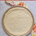 A round pizza baking pan holds freshly rolled out pizza dough, with a colorful kitchen towel partially visible beside it on a hexagon-tiled surface.
