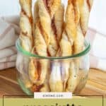 A glass jar filled with delicious cheese twists sits on a wooden surface with a striped cloth in the background.