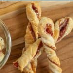 Three savory cheese twists made with prosciutto and puff pastry are arranged on a wooden cutting board, with part of a glass bowl visible on the left.