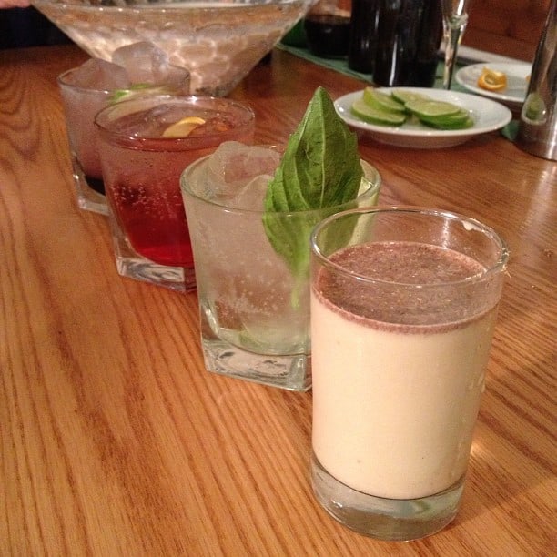 holidayPASSdrinks Four different cocktails in glass tumblers, including a refreshing basil lime spritzer, each with a unique garnish, are lined up on a wooden table next to a plate of lime slices.