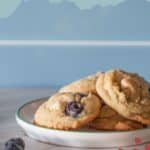 A plate of freshly baked blueberry cookies sits on a countertop with a single blueberry nearby; text above reads "Blueberry Cookies.