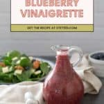 A glass bottle of blueberry vinaigrette sits on a cloth next to a fresh spinach and pecan salad. Text above reads "Blueberry Vinaigrette. Get the blueberry vinaigrette recipe at stetted.com.