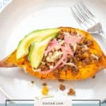 A halved baked Tex-Mex Sweet Potato topped with ground meat, pickled onions, cheese, and avocado slices is served on a white plate with a fork.