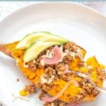 A Tex-Mex Sweet Potato half topped with ground meat, cheese, pickled onions, and sliced avocado is served on a white plate.