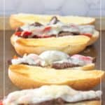Four steak & cheese sandwiches with melted cheese and red peppers on sliced hoagie rolls, arranged in a row on a baking sheet.