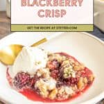 A serving of warm blackberry crisp with oats and walnuts, topped with creamy vanilla ice cream, sits on a white plate with a gold spoon.