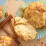 Snickerdoodle Muffins in blue paper liners, one broken open to reveal the soft inside, are displayed with cinnamon sticks on a light wooden surface.