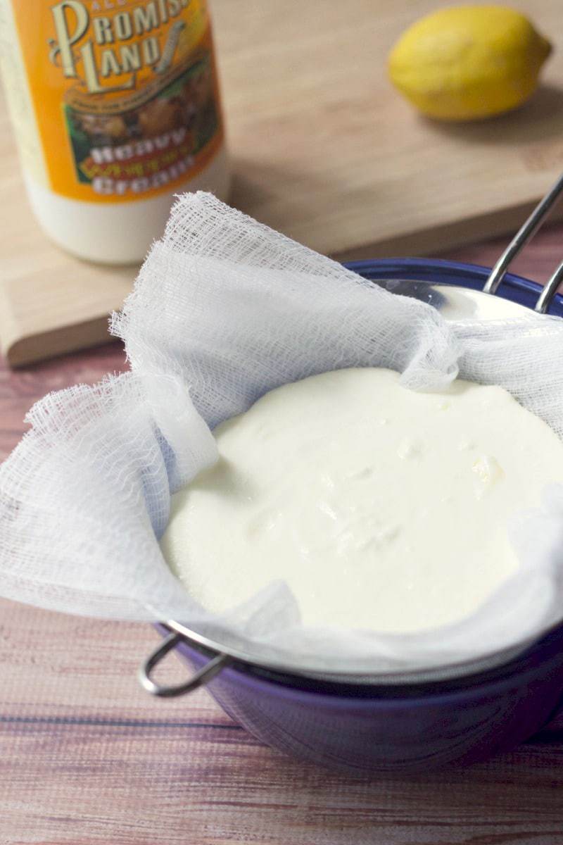 A blue bowl with a strainer and cheesecloth, with a creamy white liquid inside draining.