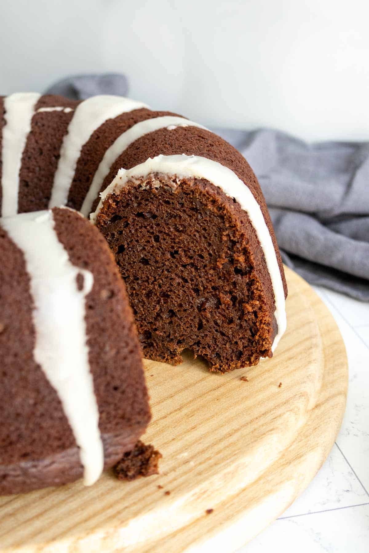 A chocolate pumpkin bundt cake with white icing drizzled on top is displayed on a wooden board, with one slice cut out.