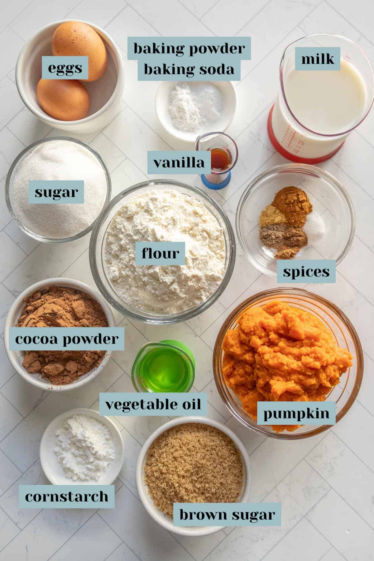 A variety of baking ingredients in bowls and cups for a chocolate pumpkin bundt, including eggs, sugar, flour, cocoa powder, pumpkin, brown sugar, spices, vanilla, baking powder, cornstarch, milk, and vegetable oil.