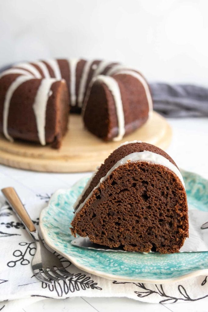A slice of chocolate pumpkin bundt cake with white icing sits on a blue patterned plate, while the rest of the cake rests on a wooden board in the background.