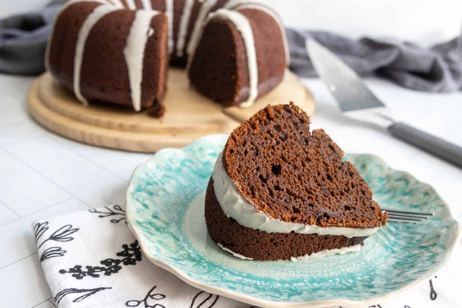 A slice of chocolate pumpkin bundt cake with white icing sits on a blue patterned plate, while the rest of the cake rests on a wooden board in the background.