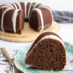 A slice of rich chocolate pumpkin bundt cake with white icing rests on a plate, while the rest of the chocolate pumpkin bundt cake sits in the background on a wooden board.