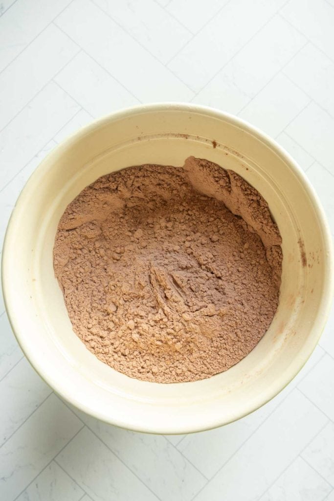 A mixing bowl containing dry cocoa powder and flour mixture for a chocolate pumpkin bundt, placed on a light tiled surface.