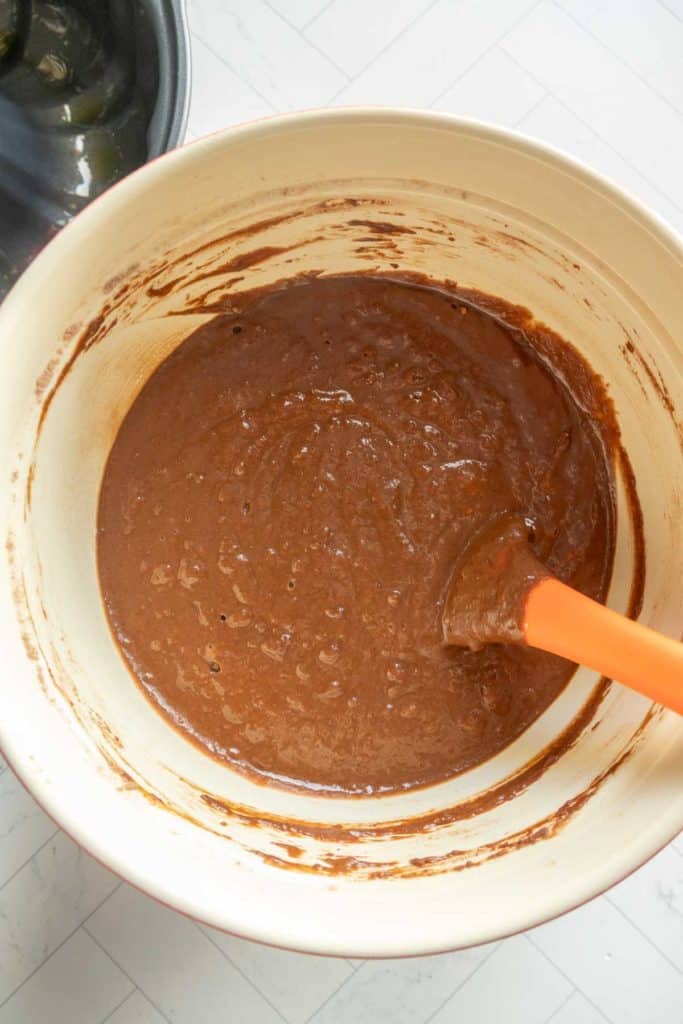 A mixing bowl filled with chocolate pumpkin bundt cake batter being stirred with an orange spatula.