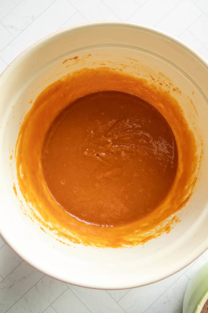 A large mixing bowl containing smooth orange batter for a chocolate pumpkin bundt, set on a light tiled surface.