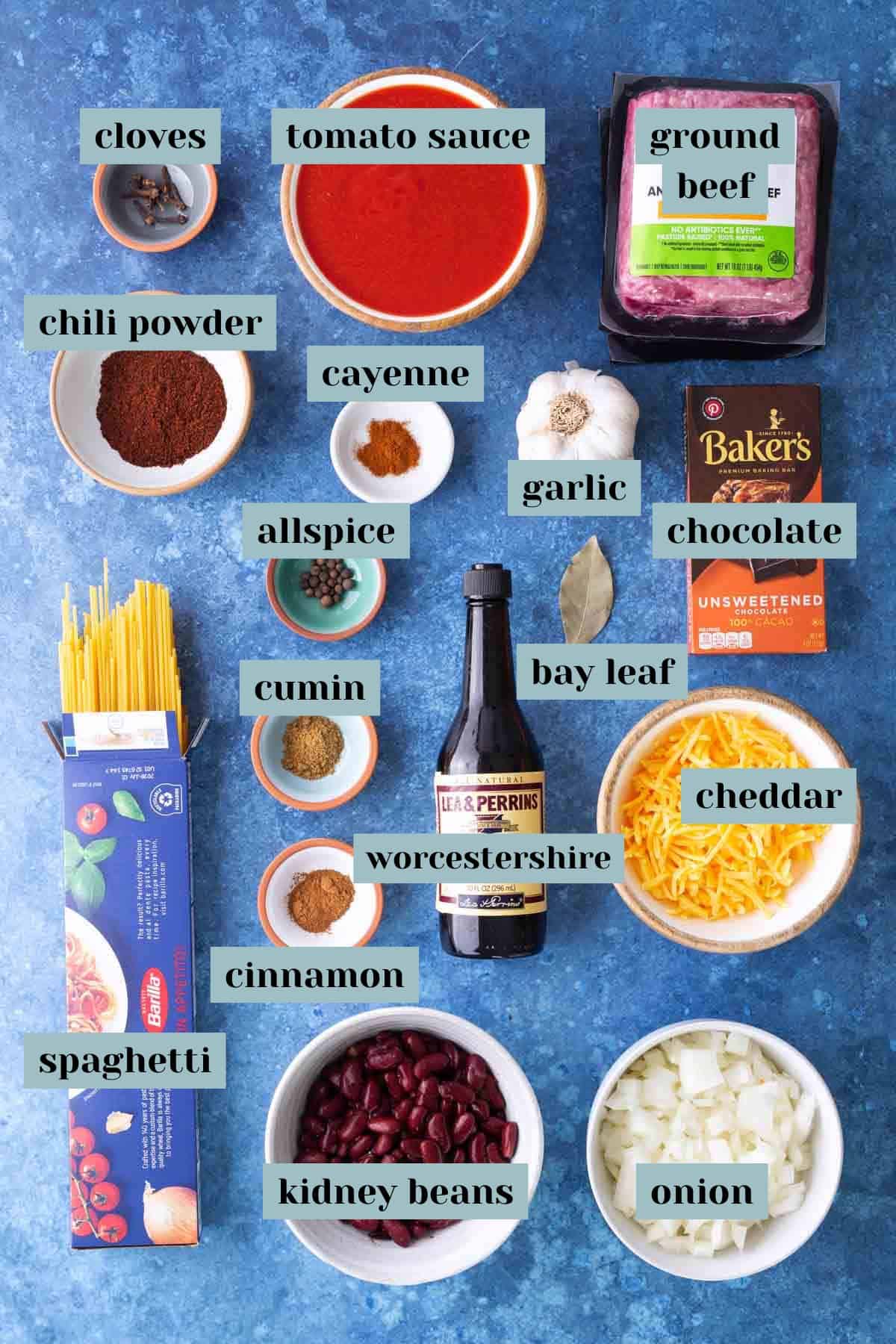 Ingredients for classic Cincinnati chili are arranged on a blue surface, featuring ground beef, tomato sauce, spices, chocolate, cheddar, spaghetti, kidney beans, onion, and garlic.