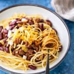 A plate of Cincinnati chili spaghetti topped with ground beef chili, kidney beans, shredded cheese, and chopped onions, with a fork resting on the side.
