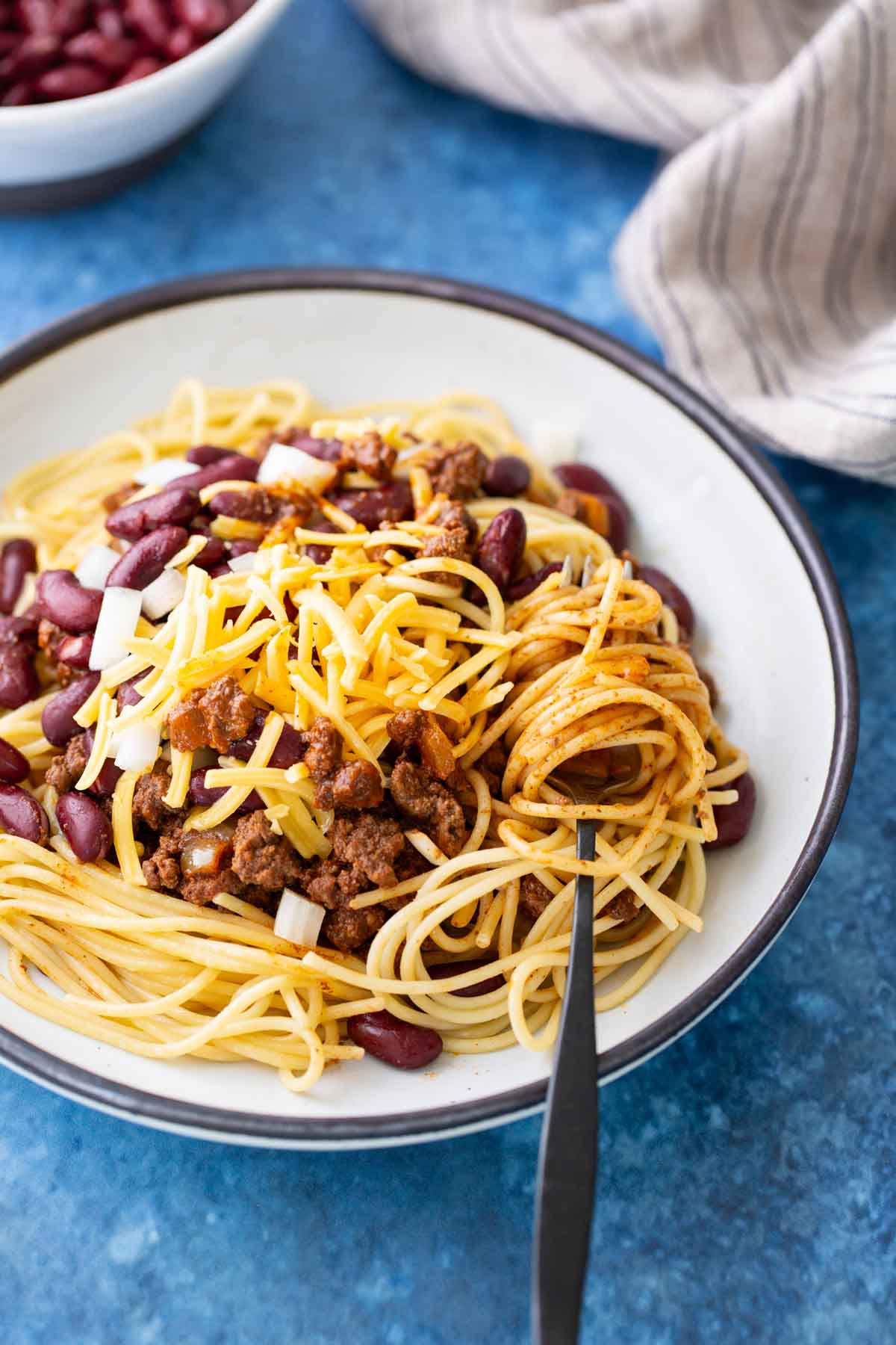 A plate of Cincinnati chili spaghetti topped with ground beef chili, kidney beans, shredded cheese, and chopped onions, with a fork resting on the side.
