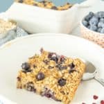 A square of blueberry baked oatmeal on a white plate with a spoon, with a dish of blueberry baked oatmeal and a bowl of fresh blueberries in the background.