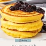 A stack of fluffy cornmeal pancakes topped with blueberry compote sits on a white plate, accompanied by syrup and more cornmeal pancakes in the background.