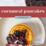 A plate with cornmeal pancakes topped with berry compote, a fork and knife, a bowl of berry sauce, and a bowl of blueberries rest on a white surface.