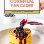 A hand spoons blueberry sauce onto a stack of fluffy cornmeal pancakes on a white plate, with a bowl of blueberries and extra sauce in the background.