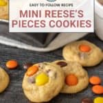 A close-up of Mini Reese's Pieces cookies on a wooden surface, showcasing the vibrant colors and delightful texture, with more delectable Reese's Pieces cookies tempting from a tray in the background.