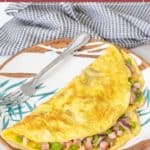 A classic Denver omelet filled with diced ham, green bell peppers, and onions is served on a plate with a fork and a checkered napkin in the background.