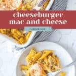 A plate and baking dish of Cheeseburger Mac and Cheese with penne pasta, ground beef, cheese, tomato slices, and breadcrumbs are served with a fork on a white surface.