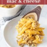 A serving of Cheeseburger Mac and Cheese on a white plate with a wooden spoon, with more creamy, cheesy goodness in a baking dish in the background.