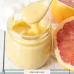 A jar of Grapefruit Curd with a spoon sits beside a halved grapefruit on a white surface.