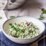 A bowl of cauliflower tabouli garnished with parsley sits on a napkin with a fork and spoon; a large bowl of cauliflower tabbouleh is in the background.