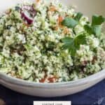 A bowl of cauliflower tabouli garnished with parsley sits on a blue napkin, featuring chopped vegetables mixed throughout the vibrant salad.