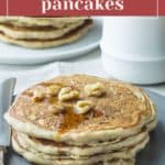 A stack of fluffy zucchini bread pancakes topped with walnuts and syrup sits on a plate with a fork; additional zucchini bread pancakes and a white jug are in the background.