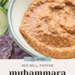 A vibrant bowl of red bell pepper muhammara sits on a plate, paired perfectly with purple tortilla chips and crisp cucumber slices.