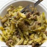 A white bowl filled with savory pork cabbage skillet—tender cooked cabbage and ground pork, with a fork picking up a bite. Text overlay reads “pork & cabbage skillet.”.