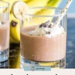Glass of chocolate smoothie topped with whipped cream and chocolate chips, garnished with a banana slice, with another glass and bananas in the background—perfect for enjoying delicious chunky monkey smoothies.