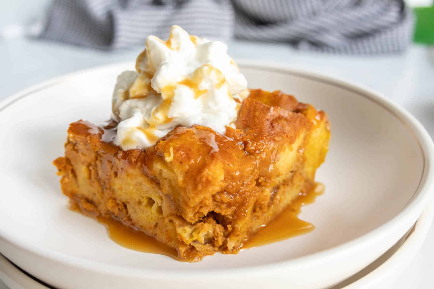 A square slice of pumpkin bread pudding topped with whipped cream and caramel sauce sits on a white plate.