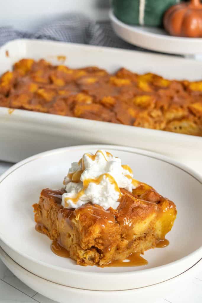 A slice of caramel-drizzled pumpkin bread pudding topped with whipped cream on a white plate, with a dish of bread pudding in the background.
