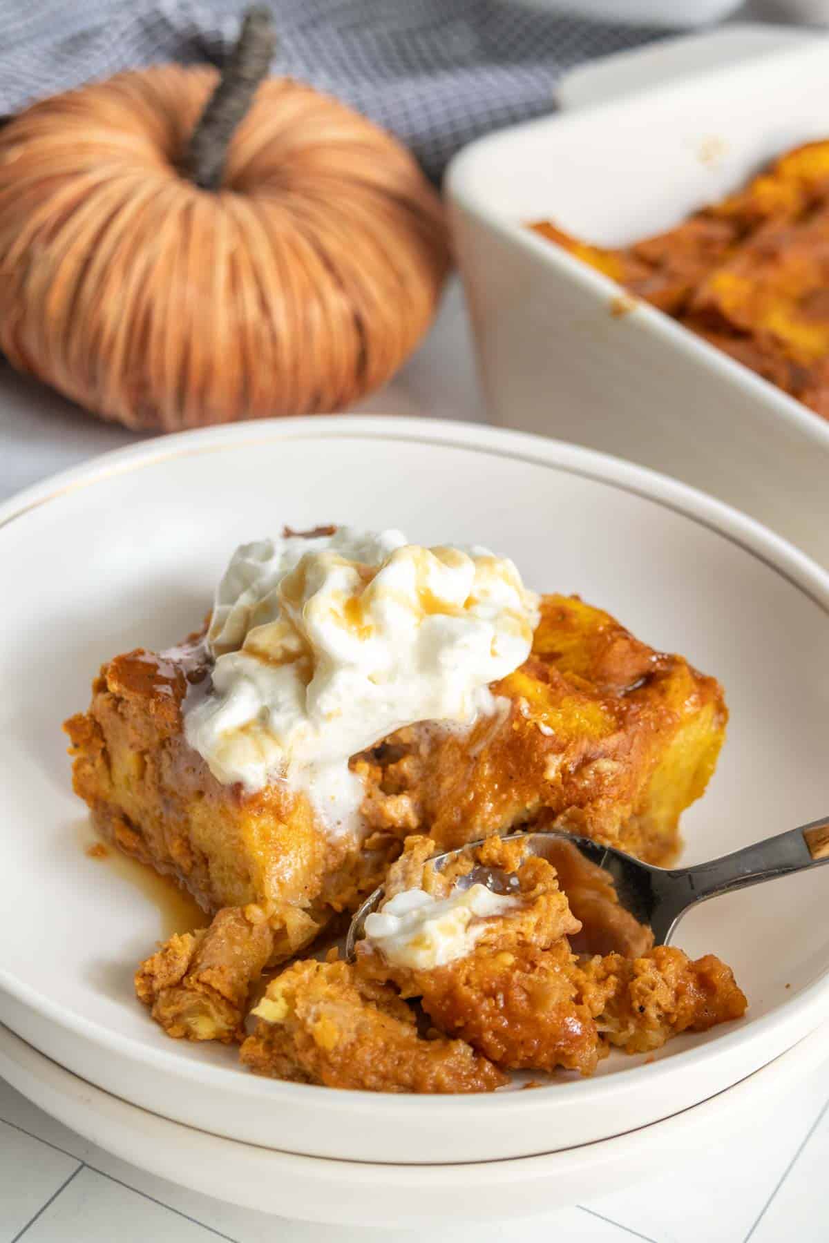 A slice of pumpkin bread pudding topped with whipped cream sits in a bowl, with a fork taking a bite; in the background, a casserole dish and decorative pumpkin complete this inviting autumn dessert scene.