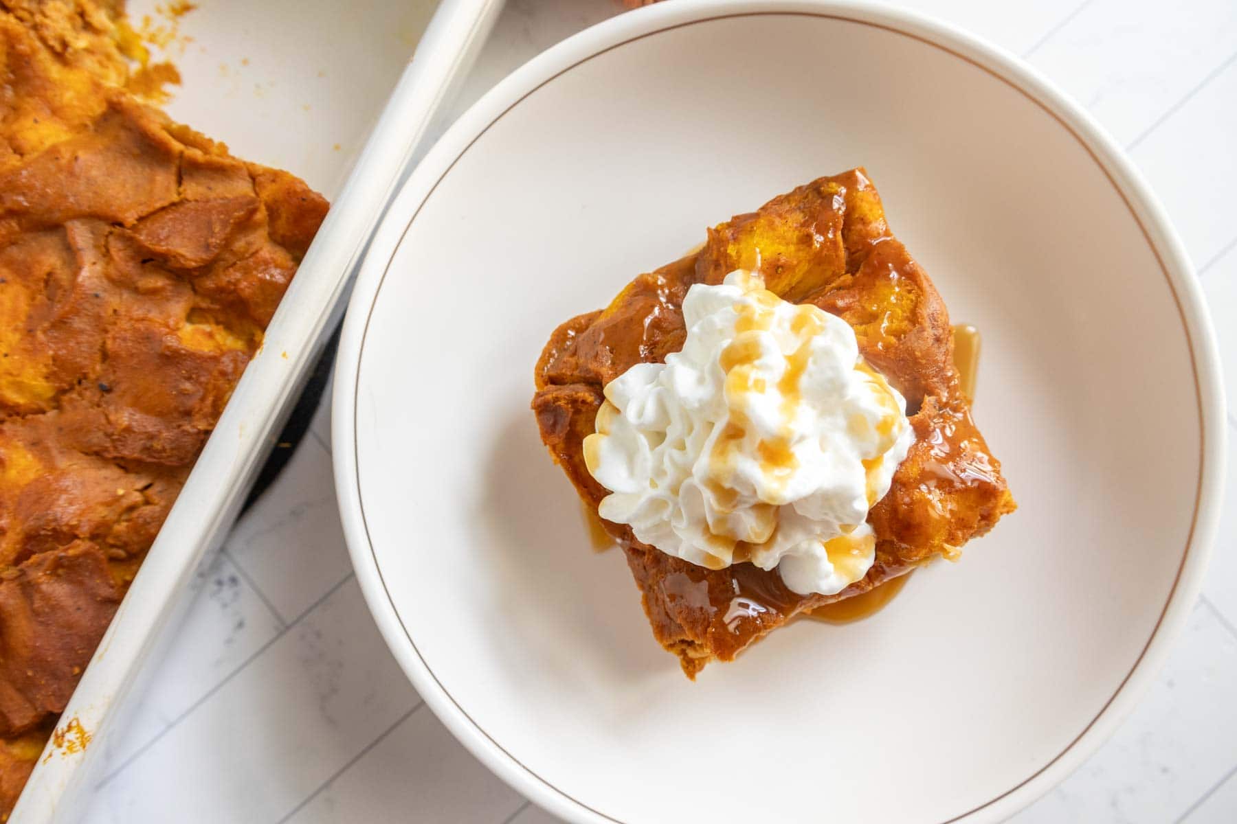 A square piece of pumpkin bread pudding topped with whipped cream and caramel sauce sits on a white plate, next to a baking dish filled with more of the decadent dessert.