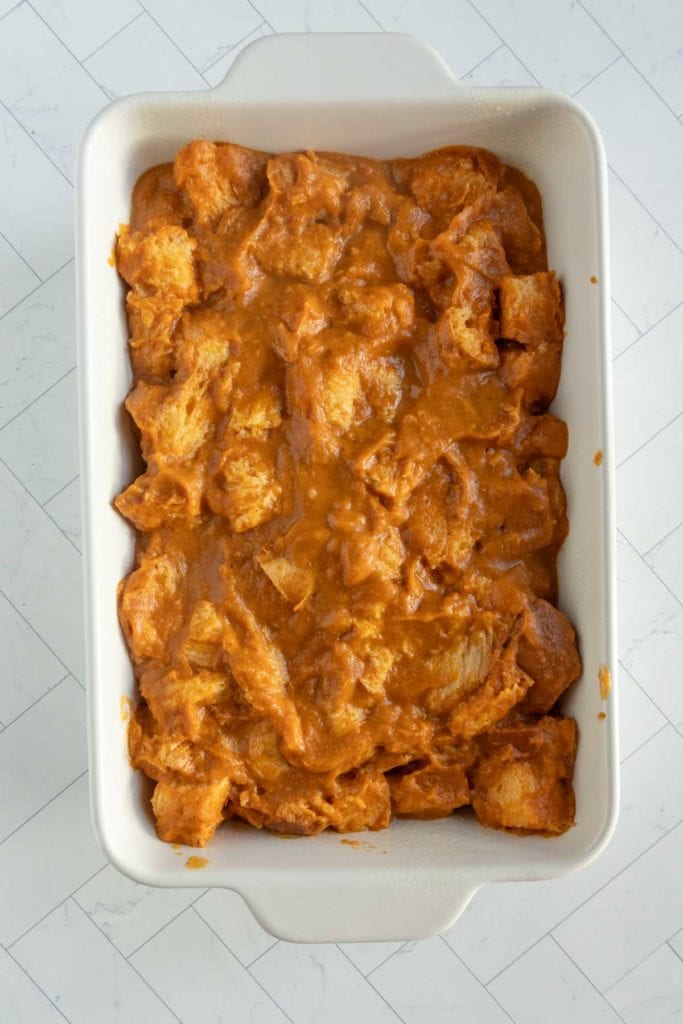 A white rectangular baking dish filled with pumpkin bread pudding, featuring bread cubes coated in an orange-brown sauce, sits on a light-colored surface.