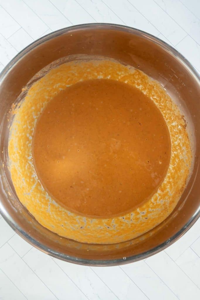 A large metal mixing bowl containing orange pumpkin bread pudding batter, with some batter residue on the sides, sits on a white tiled surface.