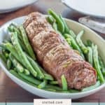 A simple steak roulade is served in an oval dish with fresh green beans, set on a wooden table with plates in the background.