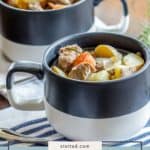 Two black and white mugs filled with hearty Irish lamb stew—brimming with tender lamb, potatoes, and carrots—sit on a table set with a striped cloth and a spoon.