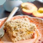 A slice of peach coffee cake with crumb topping sits on an orange patterned plate beside a fork; fresh peach halves are in the background, highlighting this inviting peach coffee cake treat.