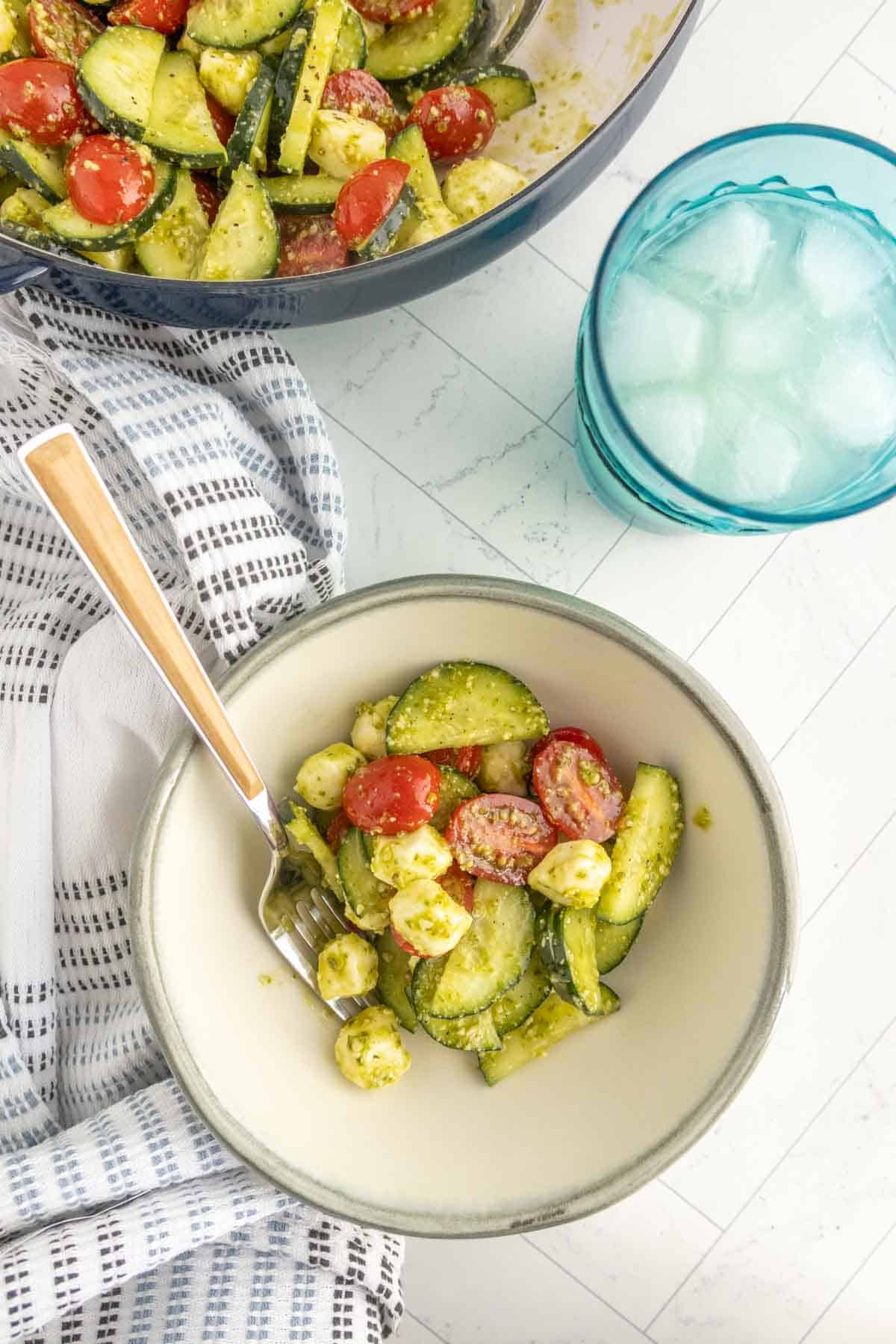A bowl of cucumber tomato salad with mozzarella balls and pesto sits next to a fork and a glass of iced water on a white surface.