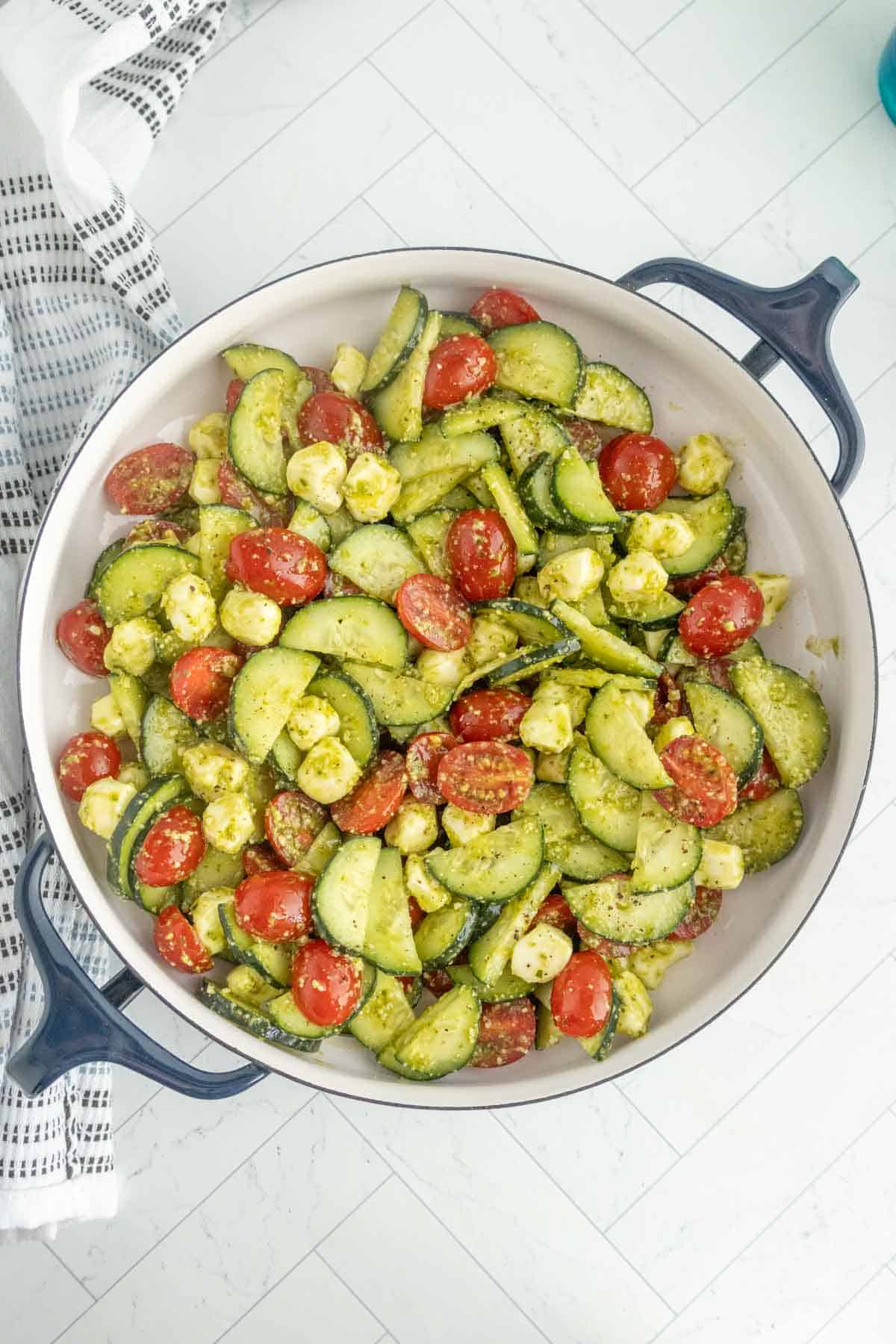 A large white bowl filled with a cucumber tomato salad featuring sliced cucumbers, cherry tomatoes, and small mozzarella balls, all lightly coated in a green herb dressing.